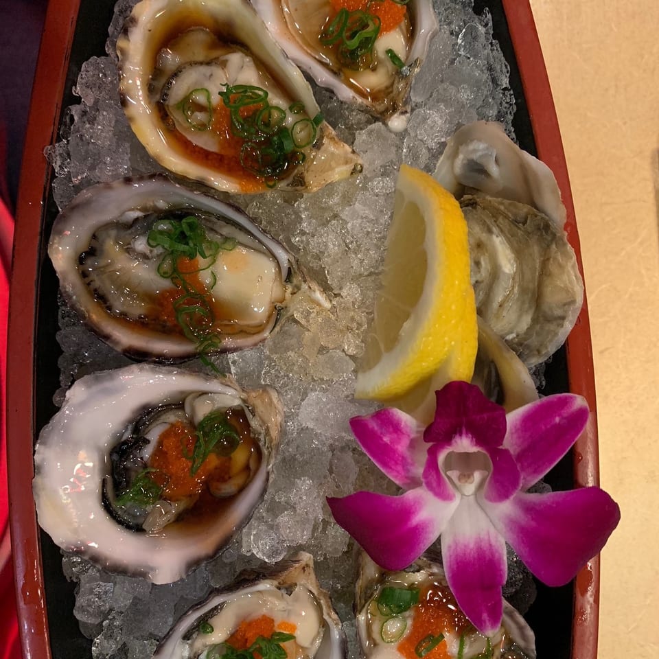 Six oysters on the half shell with roe and scallions, a lemon wedge, and purple orchid on ice.