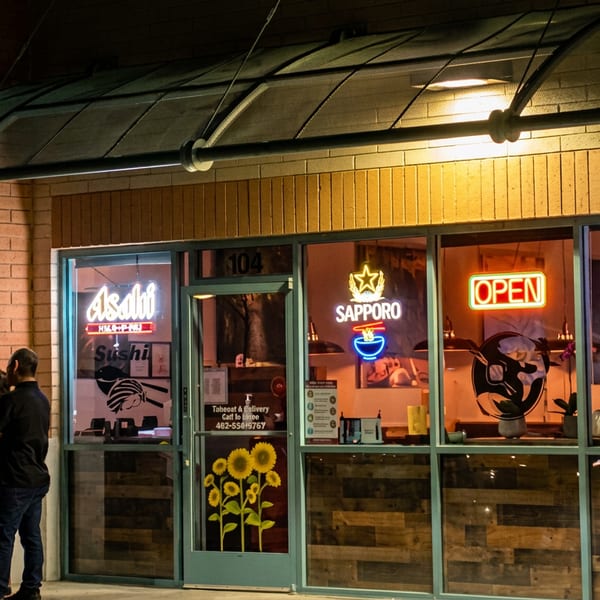 Exterior of Asahi Sushi restaurant at night with neon signs and people standing outside.