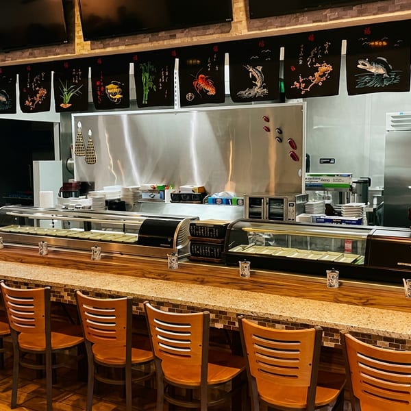 Empty sushi bar with wooden stools, display cases, and decorative banners above a stainless steel kitchen.