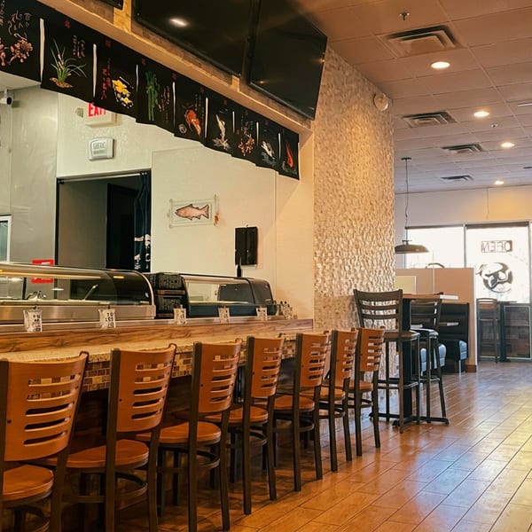 A man walks past a sushi bar with wooden chairs and a display case, towards dining tables.